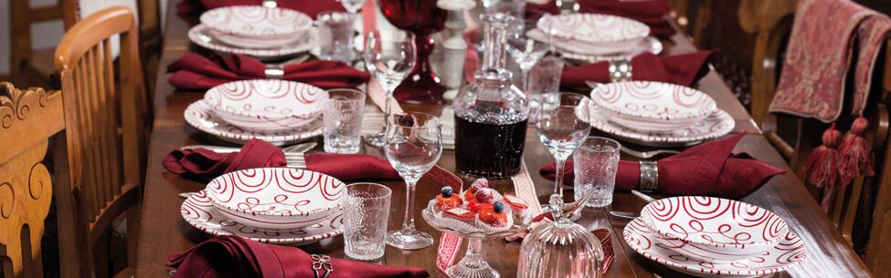 Festlich gedeckter Holztisch mit weißen Tellern, Kristallgläsern, roten Servietten und einer zentralen Blumendekoration.